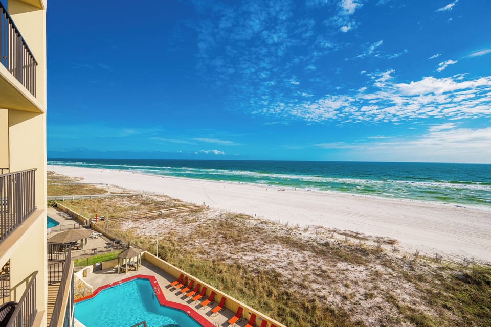 The view showcases the vibrant pool area with lounge chairs and a stunning beachfront, framed by white sand and blue waters