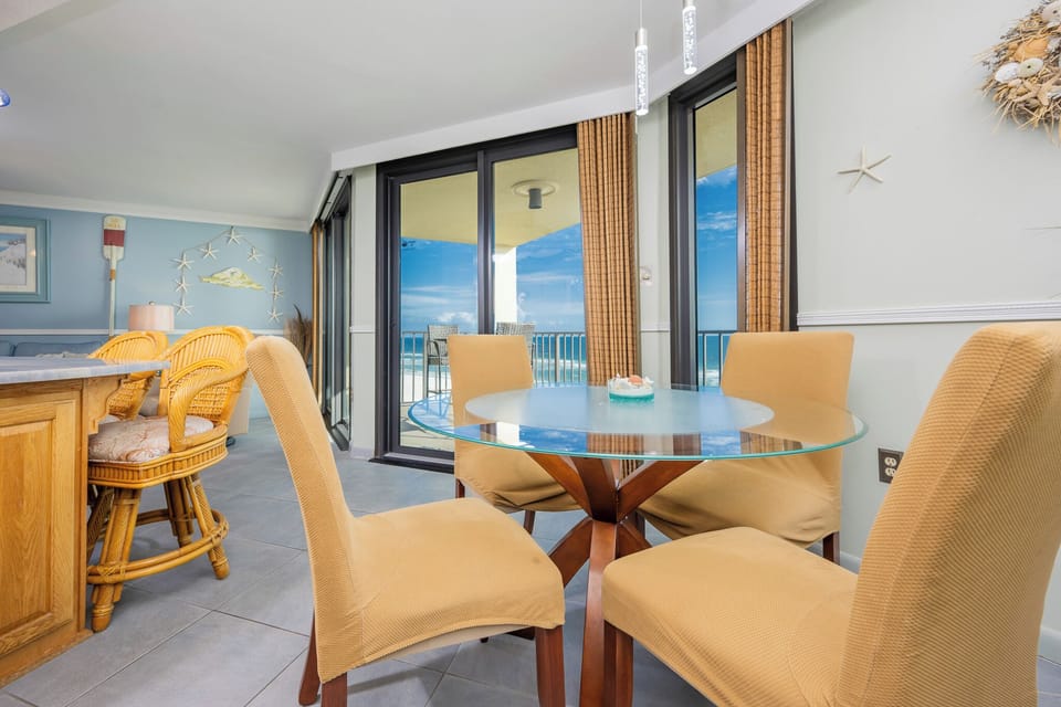 A glass-top dining table surrounded by four beige chairs offers a view of the beach through large sliding glass doors