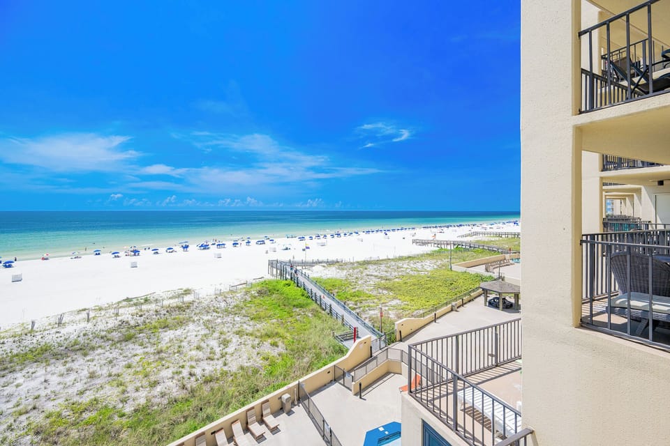 Vibrant beach view with white sand and blue umbrellas, framed by a clear blue sky and lush greenery