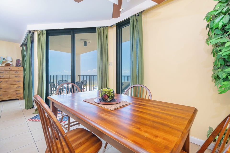 A dining area features a wooden table with four chairs, a potted plant, and large windows showcasing the beach front