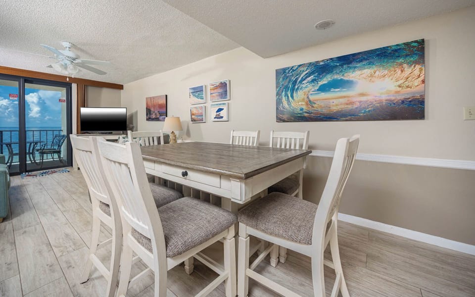 The living/dining area features a large table surrounded by white chairs, beach artwork, a TV, and queen sofa-sleeper