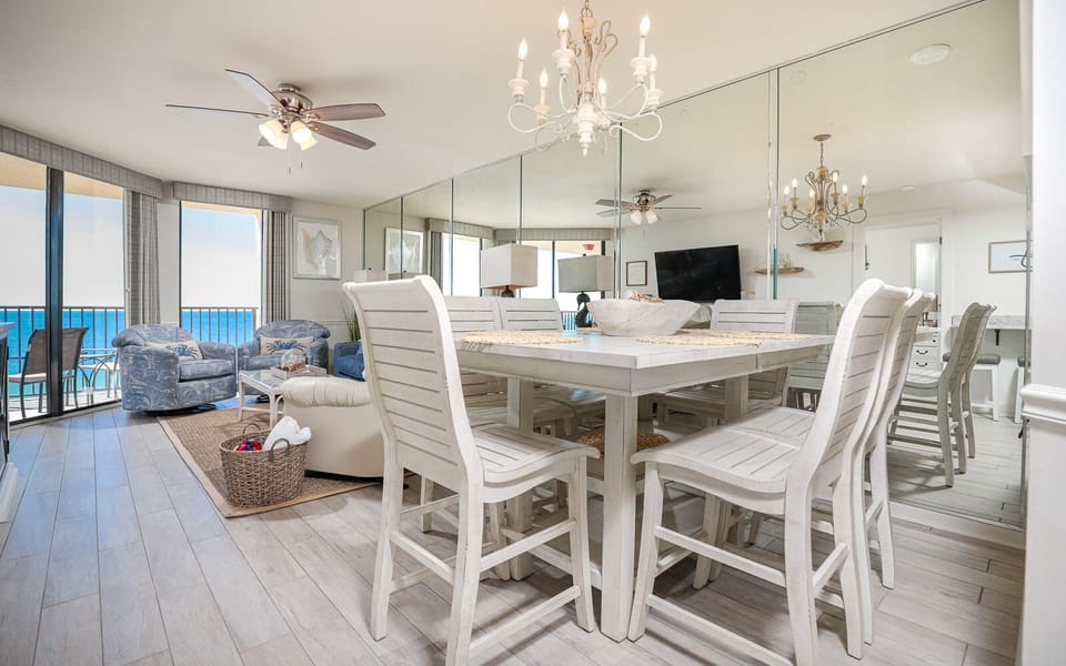 Bright dining area with white wooden furniture, a chandelier, and views of the beach through large windows