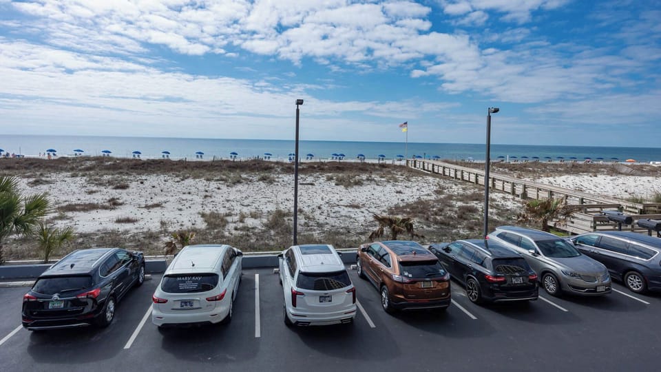 A view of the beach with blue umbrellas and a wooden walkway, framed by parked cars in a well-maintained lot