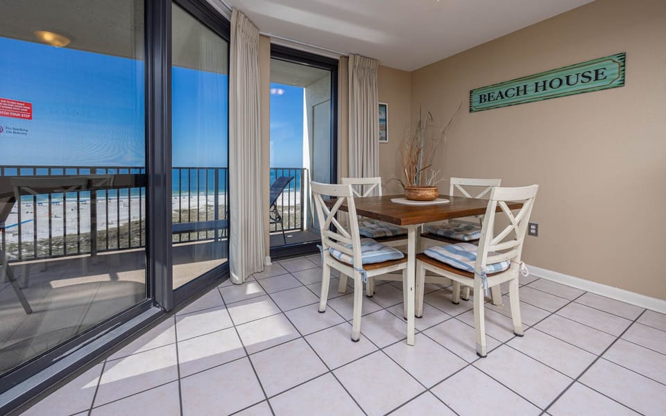 Dining area features a wooden table with cushioned chairs, offering a view of the beach through large glass doors