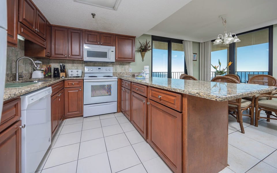 The kitchen features rich wood cabinetry, granite countertops, and a breakfast bar with views of the beach