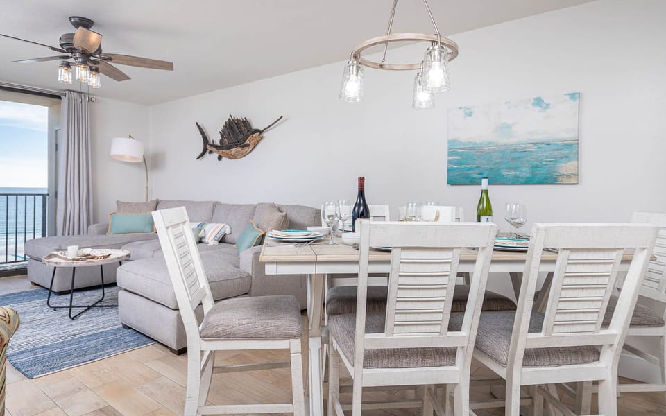 Cozy dining area features a light wood table set for six, with a view of the beach visible through sliding glass doors