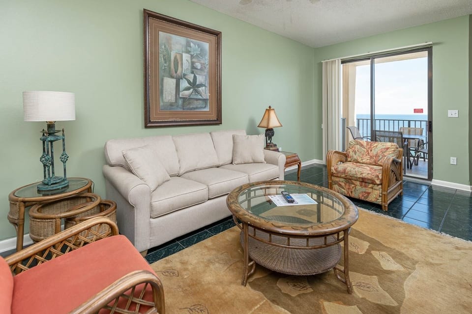A light-filled living area features a beige sofa with queen sleeper, armchair, and balcony doors leading to a beach view