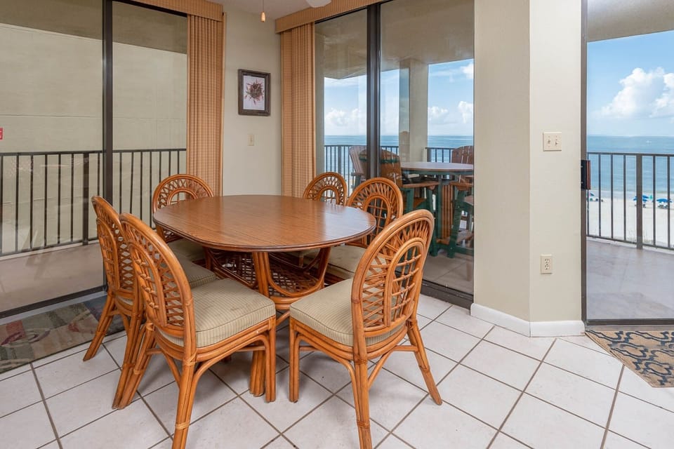 Dining area features a round wooden table surrounded by six rattan chairs, with a view of the beach