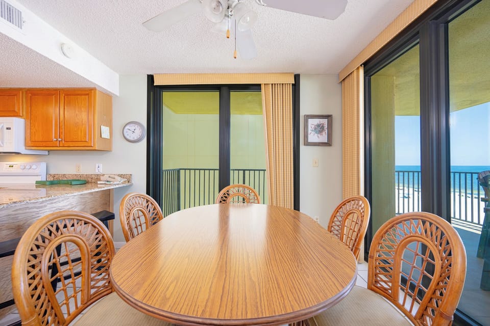 Dining area features a round wooden table surrounded by six rattan chairs, with a view of the beach