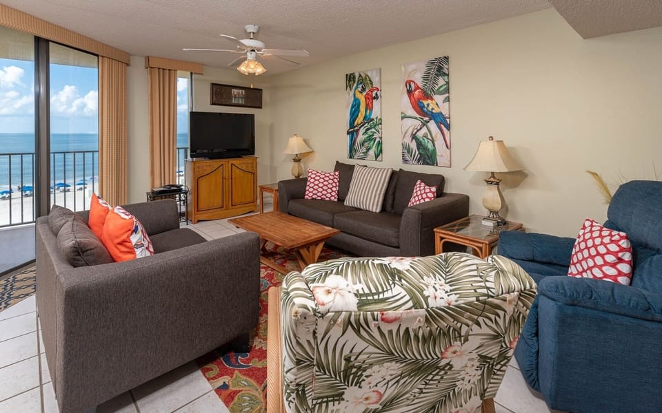 Living area features a cozy sofa with colorful throw pillows, tropical artwork, and a view of the beach through large windows