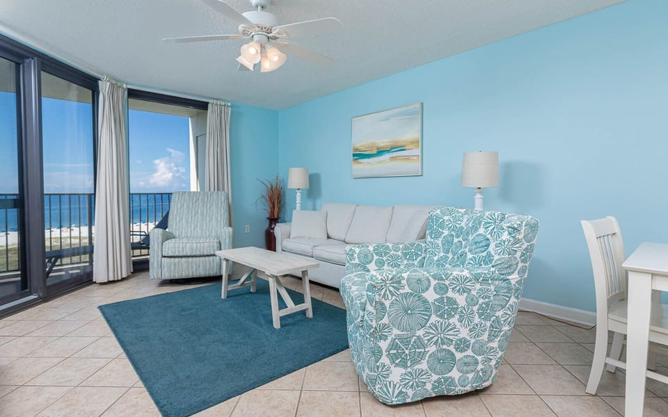 Living area features a cozy sofa with queen sleeper, patterned chairs, and a table, with a view of the beach from the balcony