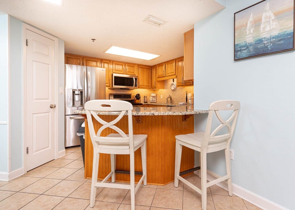 Kitchen area features wooden cabinetry, granite countertops, and a breakfast bar with two white stools for casual dining