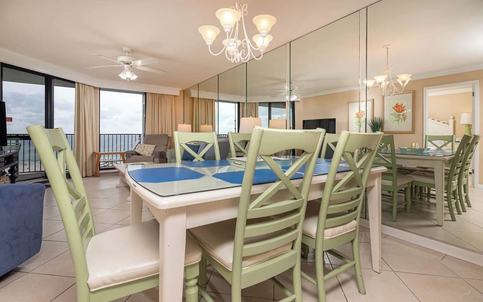 Dining area features a glass-top table with green chairs, overlooking the beach through large windows and soft curtains
