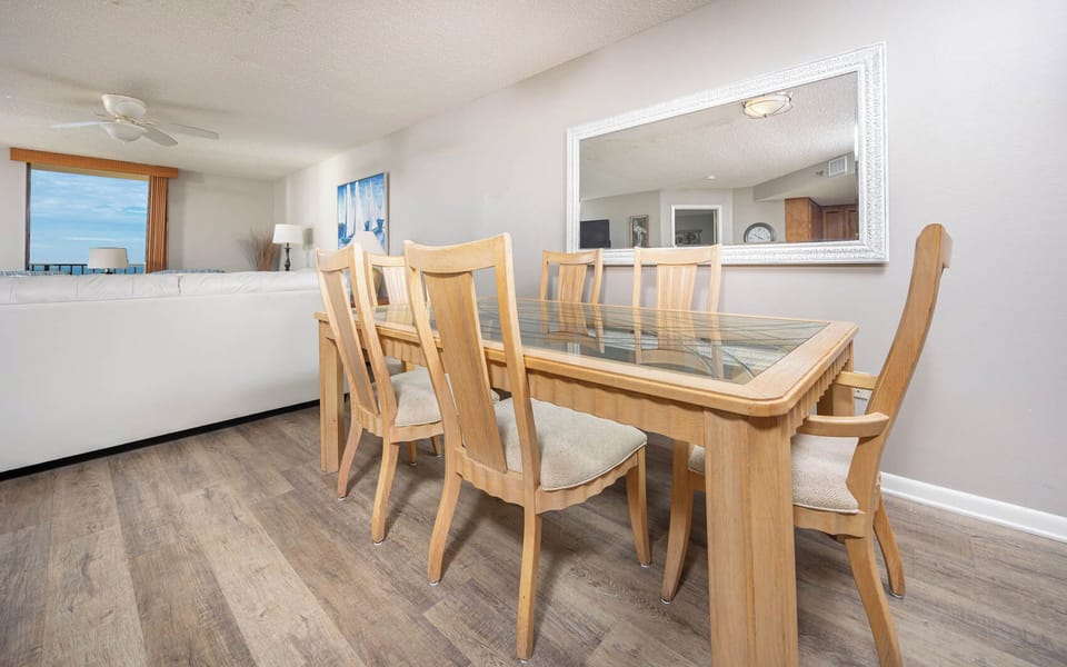 A wooden dining table with glass top and beige chairs is set against a backdrop of a bright living area and beach view