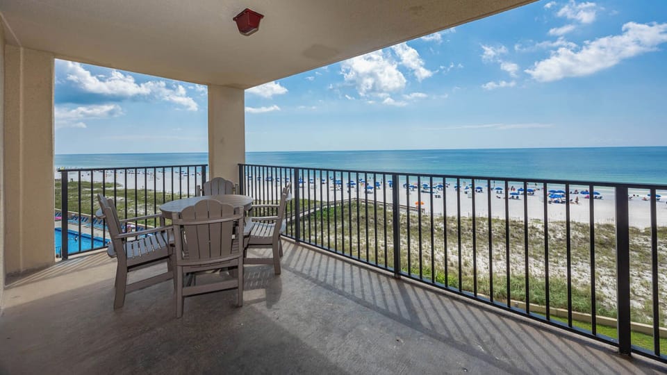 The balcony features a round table with chairs, offering a serene view of the beach and colorful umbrellas on the shore