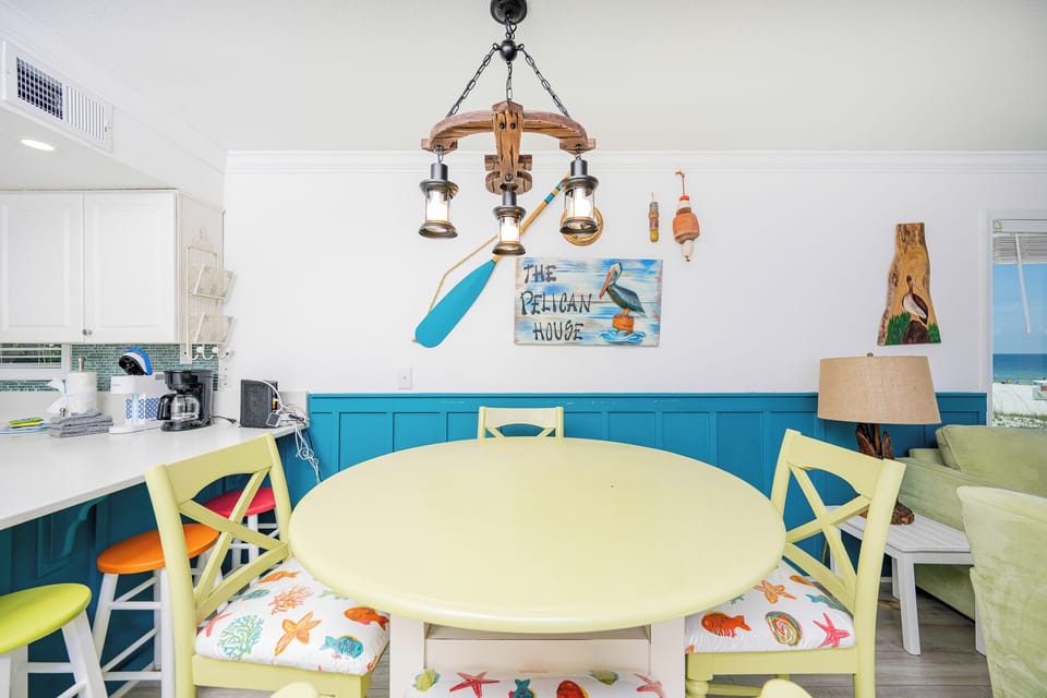 This cheerful dining area features a bright yellow table, colorful chairs, and a view of the beach, perfect for family meals