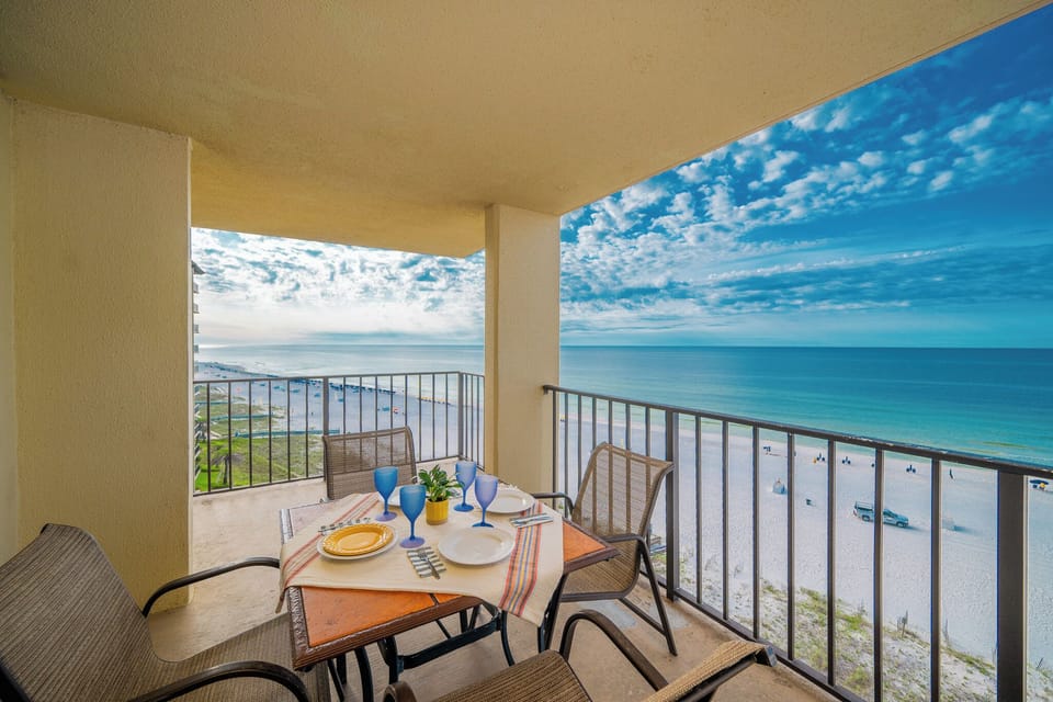 A dining table set for six overlooks a tranquil beachfront, with blue skies and gentle waves visible in the distance