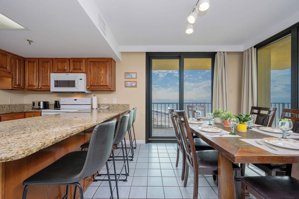 The kitchen features wooden cabinetry, a granite countertop, and a dining area with a view of the beach through large windows