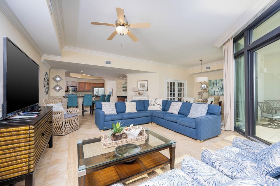The living area features a large blue sectional sofa, a TV, and opens to a bright balcony with a beach view