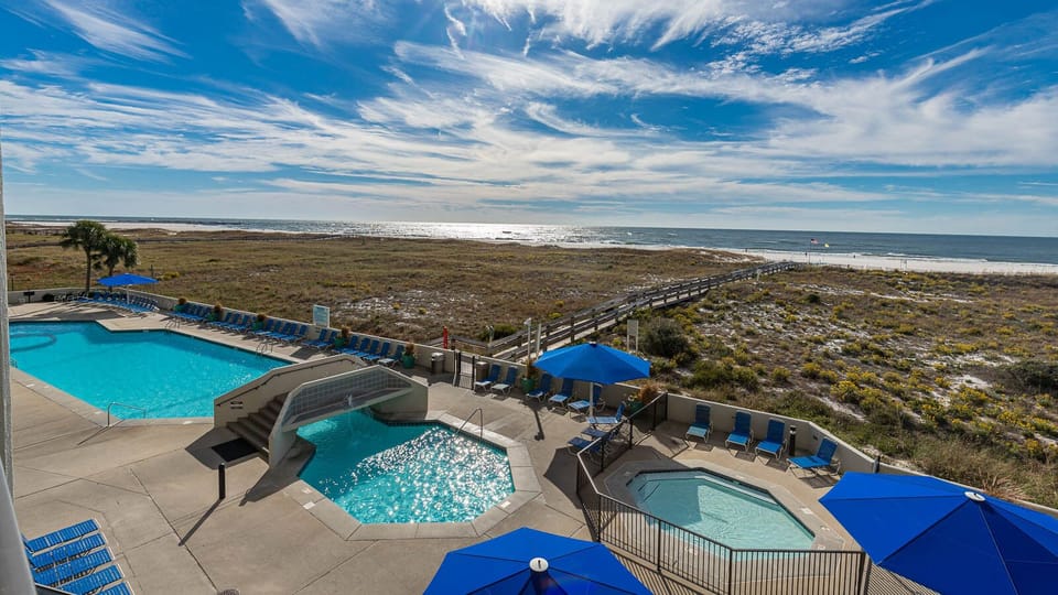 Inviting pool area features a sparkling pool and kiddie pool, with lounge chairs and a scenic view of the beach and clear sky
