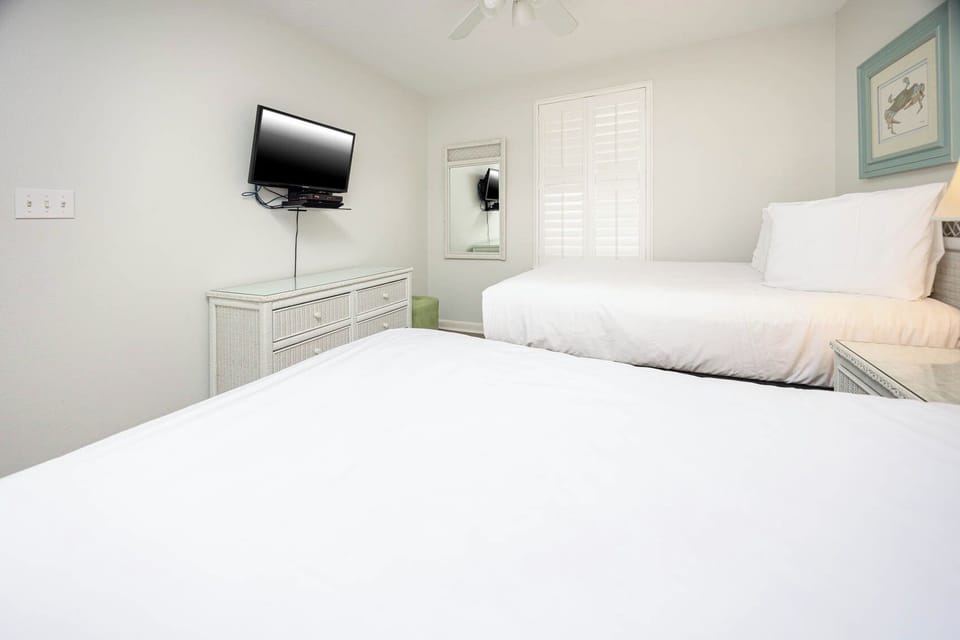 Bright guest bedroom featuring two double beds, a wall-mounted TV, and a dresser, complemented by natural light