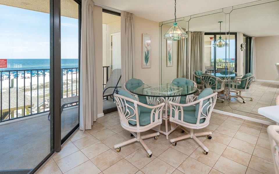 The dining area features a glass table surrounded by light-colored chairs, with a view of the beach