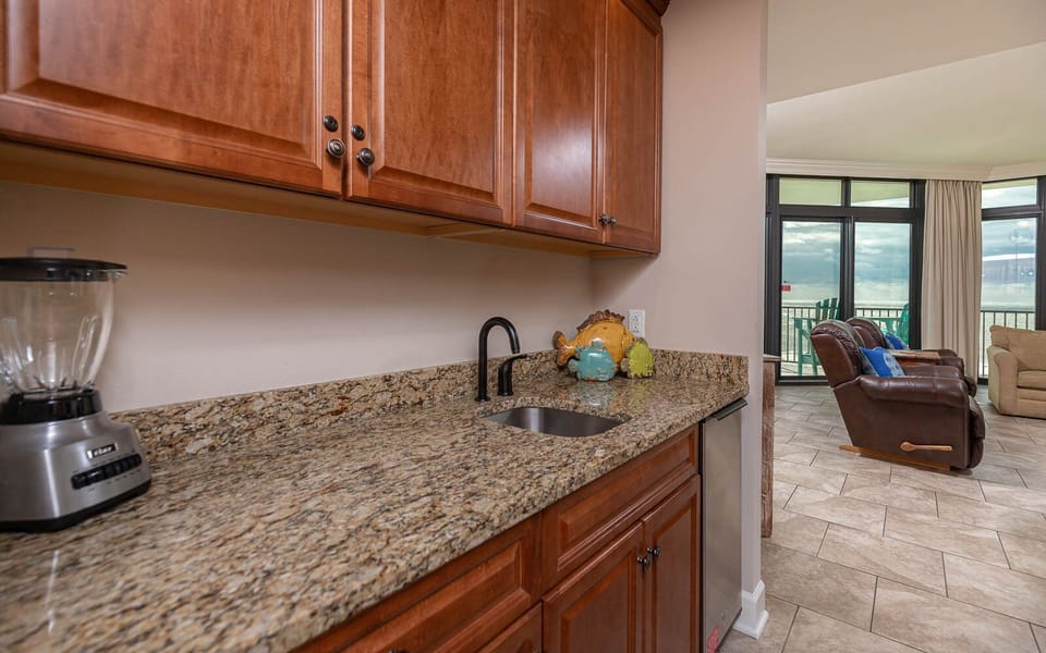 This wet bar features rich wooden cabinetry, a sleek granite countertop, and a cozy view of the inviting living area