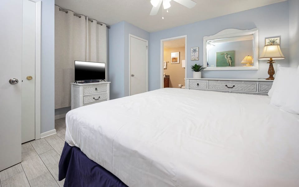 Bright guest bedroom featuring a large king bed, a TV on a decorative dresser, and a doorway leading to a bathroom