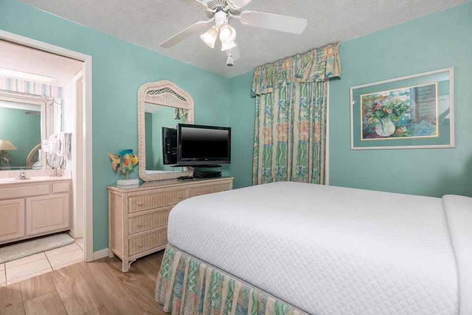 Turquoise walls complement a cozy guest bedroom featuring a queen bed, dresser, TV, and a view into the adjoining bathroom