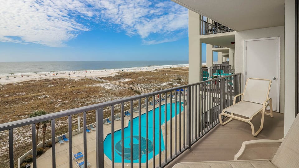 Balcony view overlooking the pool area and the beach, featuring white sand and gentle waves under a bright blue sky