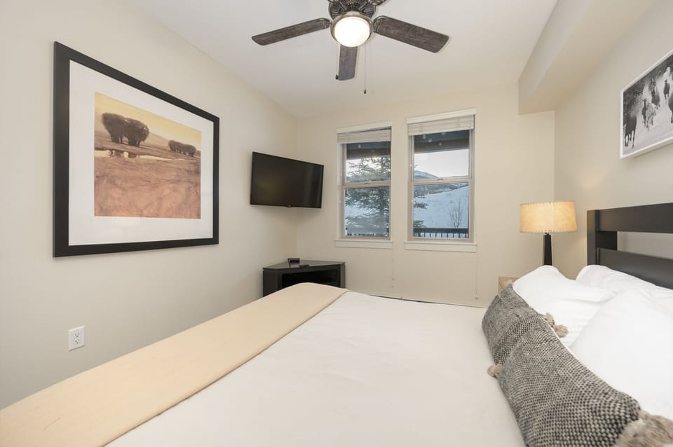 - Comfy bed faces a wall mounted TV for relaxing movie nights from under the covers - Natural light streams through twin windows showcasing outdoor views - Ceiling fan, nightstand lamp, and framed art complete the cozy setting