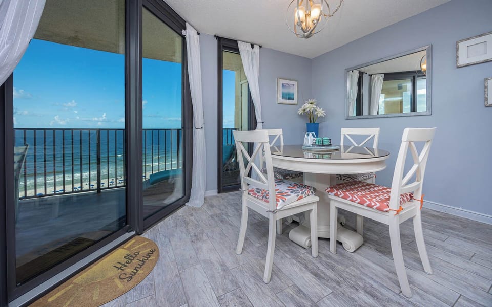 Dining area with a round table, colorful cushioned chairs, and sliding glass doors revealing a stunning beachfront view