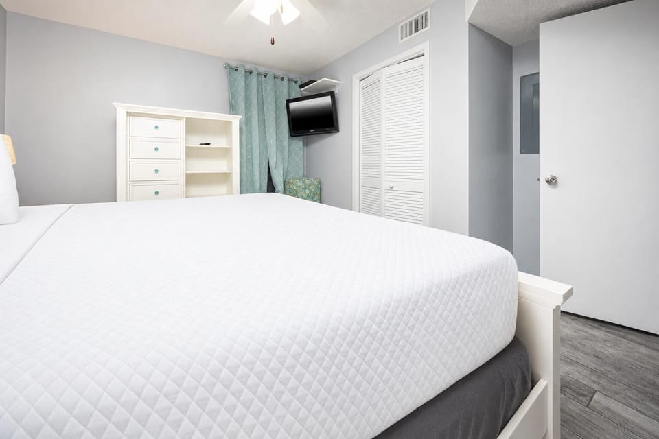 Bedroom featuring a king bed, teal curtains, a TV mounted on the wall, and a stylish dresser for storage