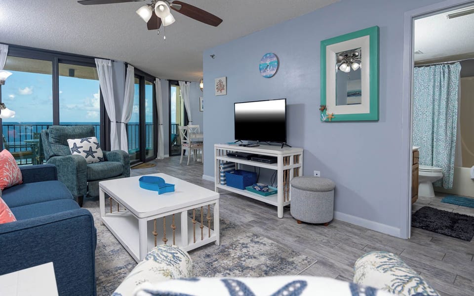 Bright living area features a cozy blue sofa, a TV stand, and large windows showcasing a serene beach front view
