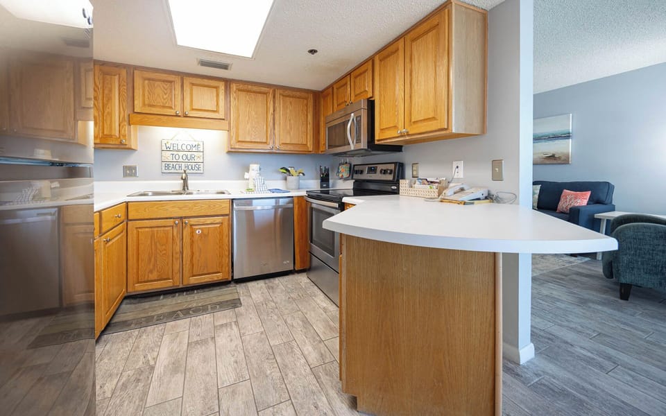 Wooden cabinetry and stainless appliances complement this kitchen, featuring a breakfast bar and a welcoming beach house sign