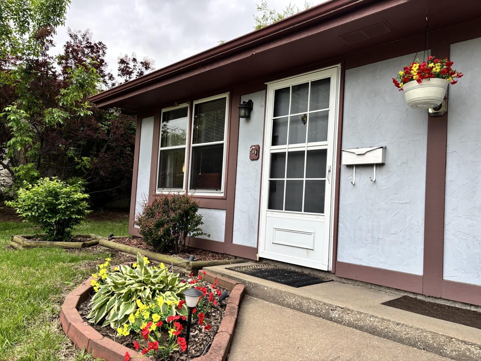 Front entrance features a white screen door, mauve trim, and a flowerbed with yellow and red blooms.