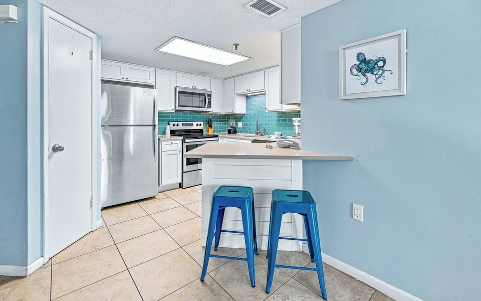 Bright kitchen featuring stainless appliances, teal backsplash, and two turquoise stools at the counter for casual dining