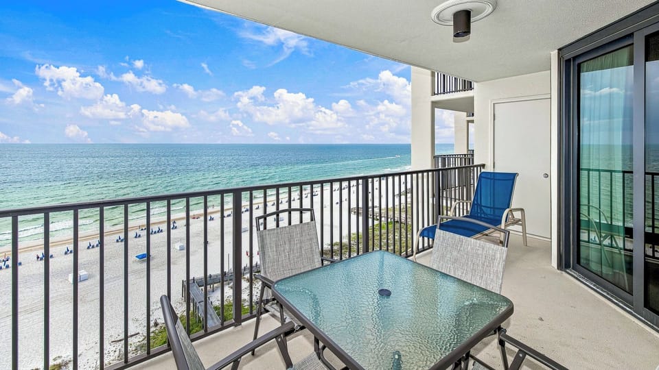 A stylish balcony features a glass-top table and six chairs, offering panoramic views of the beach and vibrant umbrellas