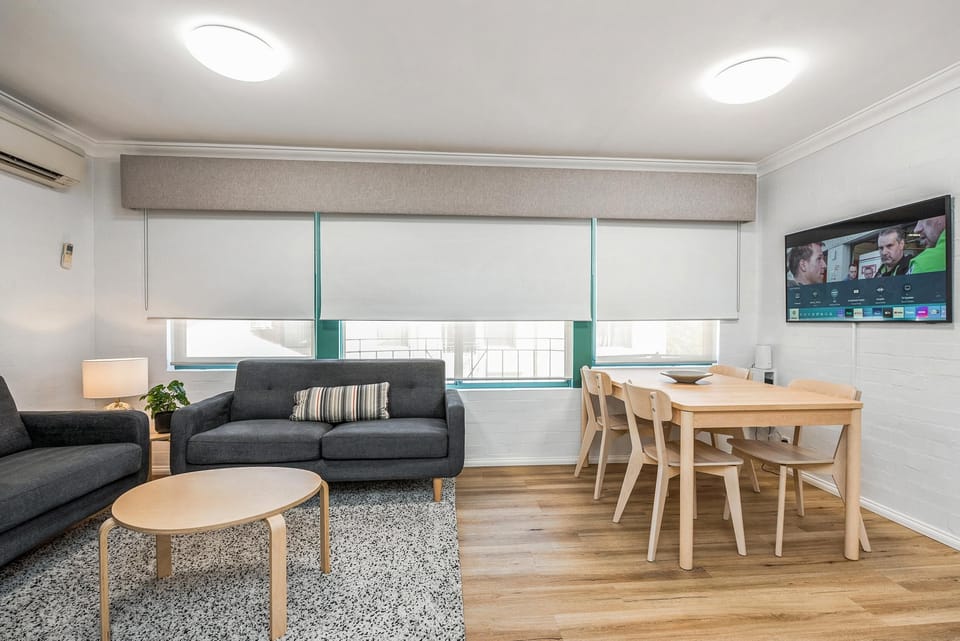 Living area with charcoal sofas, wood dining set for four, wall-mounted TV, and light wood flooring.