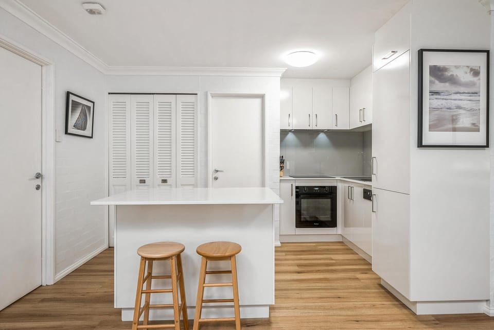 Kitchen with white cabinetry, gray glass backsplash, built-in oven, and island seating with two wooden stools.