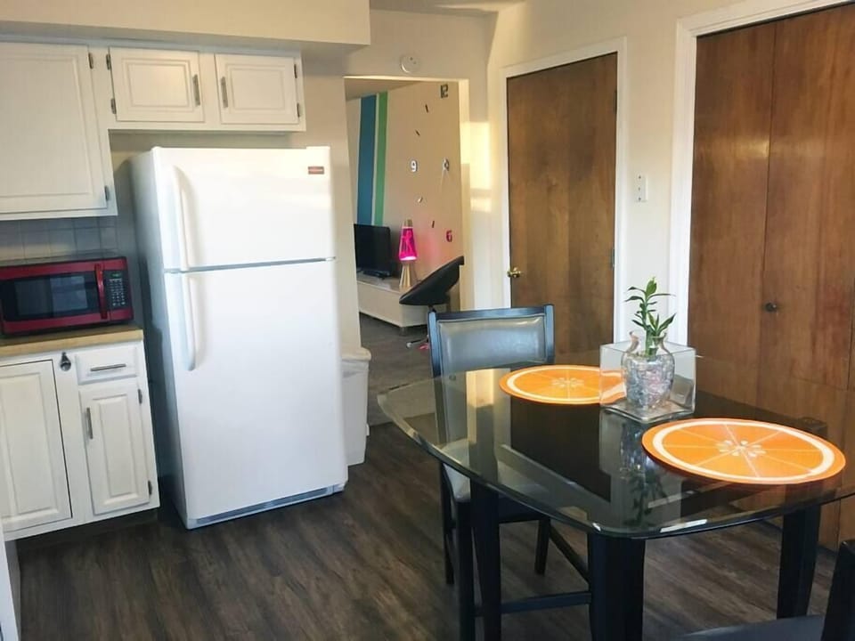 Glass-top dining table with black chairs sits on dark wood floors near white cabinetry and a white refrigerator.