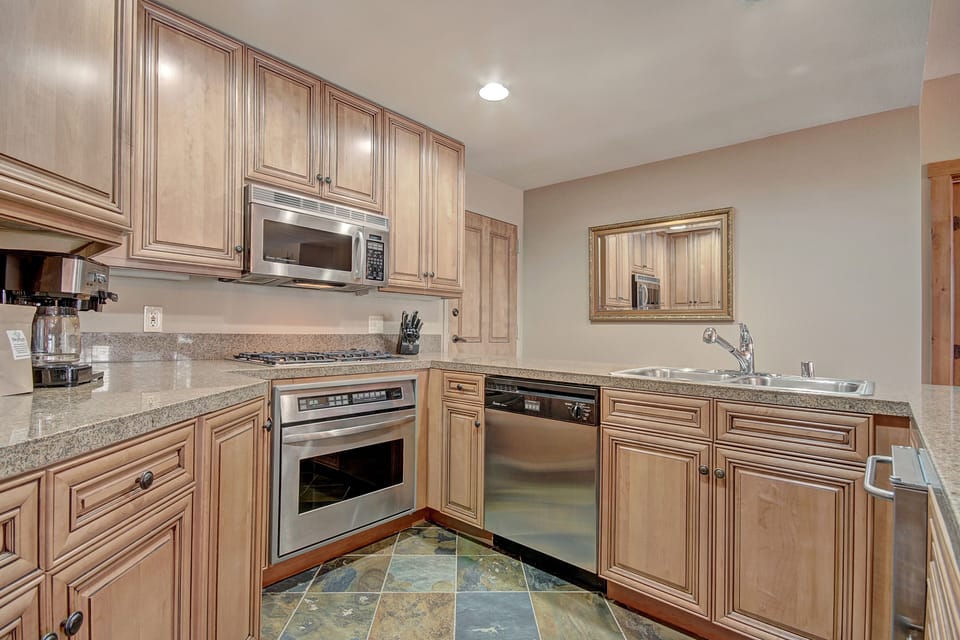 A modern kitchen with wooden cabinets, stainless steel appliances including a microwave, oven, and dishwasher, granite countertops, and a tiled floor. A mirror hangs above the sink.