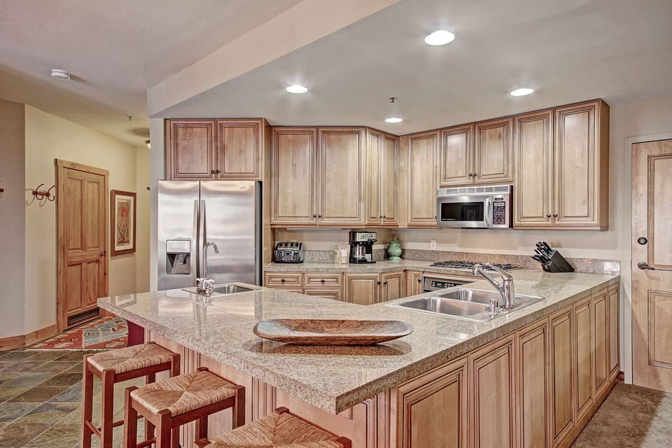 A modern kitchen with wooden cabinets, stainless steel appliances, granite countertops, an island with a sink, and three bar stools. Recessed lighting illuminates the space.