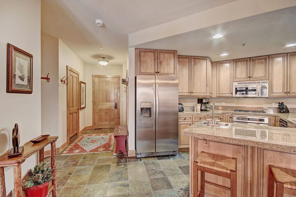 A kitchen with wooden cabinets, stainless steel appliances, a granite countertop island with seating, a red bench, and a hallway leading to a wooden door.