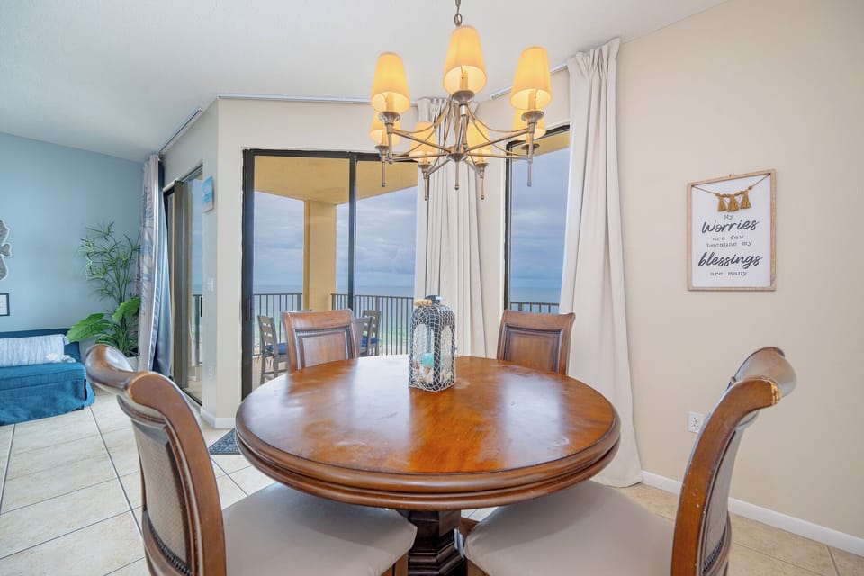 A round wooden dining table with cushioned chairs is illuminated by a stylish chandelier, offering a view of the beach