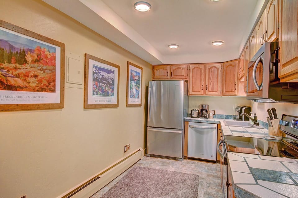 A small kitchen with wooden cabinets, stainless steel appliances, tile countertops, and framed art on the wall. The room is well-lit with ceiling lights.