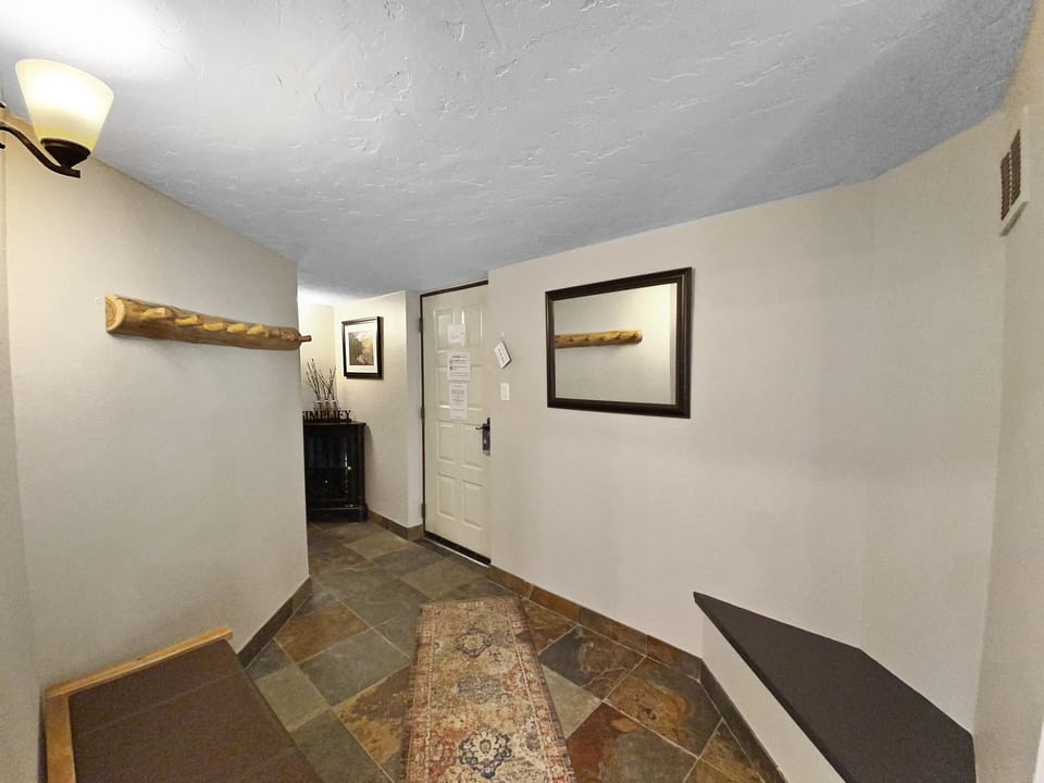 A small entryway featuring a bench, a rug, a wall-mounted coat rack, and a framed mirror. The door has several papers attached, and a side table with decor items is visible in the corner.