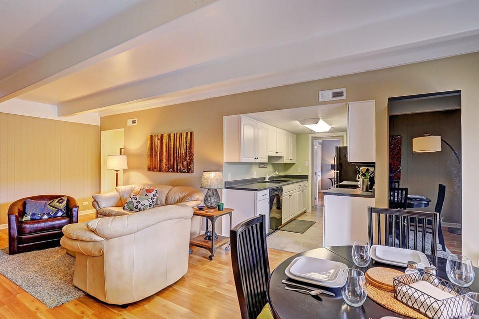 A well-lit, modern open-concept living and dining area with beige sofa, an armchair, and a dining table set for four. The kitchen is visible in the background with white cabinets and stainless steel appliances.