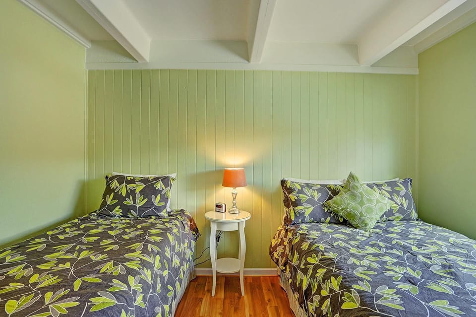 A small bedroom with two single beds, green leaf-patterned bedding, a white side table with a lamp, and light green walls.