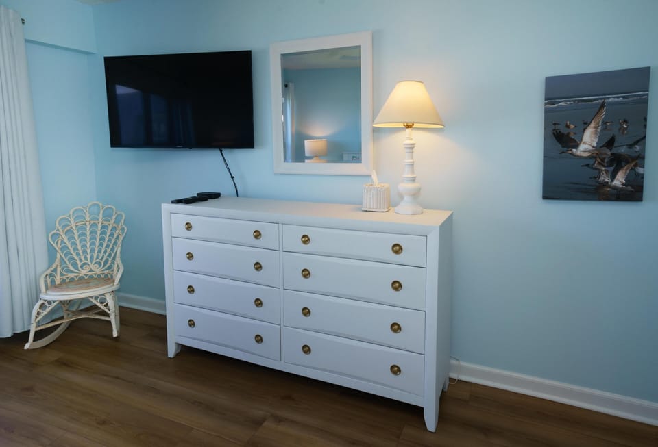 Second bedroom features pale blue walls, white dresser with brass knobs, wall-mounted TV, and wicker chair.